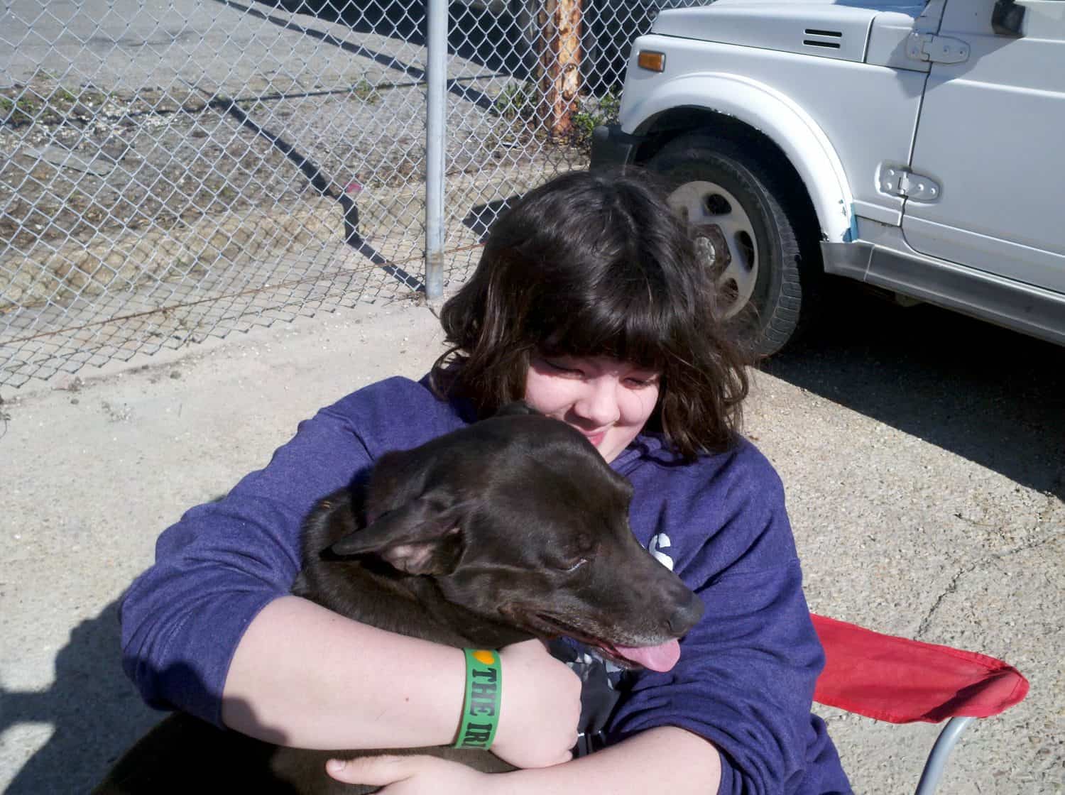 A person with dark hair hugs a large brown dog while sitting outside near a silver vehicle and chain-link fence. The person is smiling, and the dog has its tongue out.