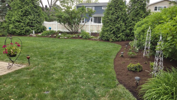 A well-maintained backyard with a green lawn, garden beds along the fence, tall trees, a white picket fence, small plants, and decorative structures. A patio corner with flowers is visible on the left.