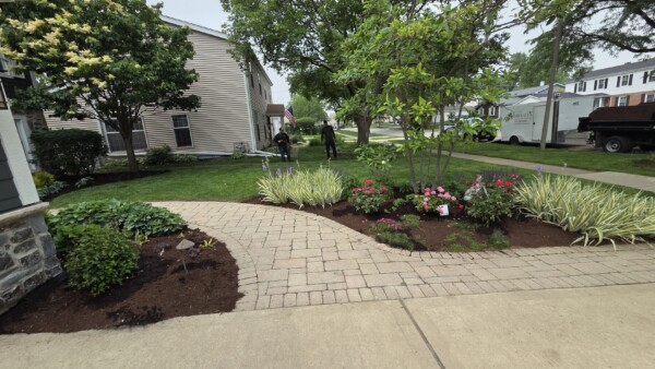 A neatly landscaped front yard with a curved stone walkway, colorful flowers, trimmed bushes, and two people gardening near the lawn. Trees and houses are visible in the background.