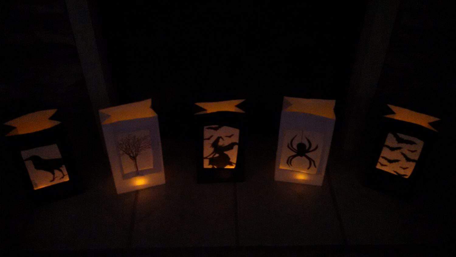 Five Halloween lanterns with silhouettes of a dog, a tree, a witch on a broom, a spider, and flying bats, all lit with a warm, yellow-orange glow in a dark setting.