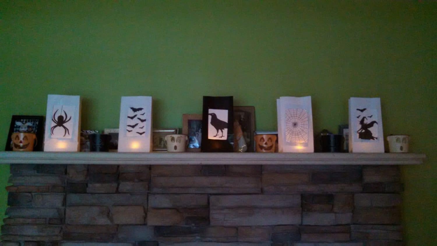 Paper lanterns with Halloween designs (spider, bats, raven, spider web, witch) sit on a stone fireplace mantel, glowing with candlelight. Small pumpkins and mugs are also displayed against a green wall.