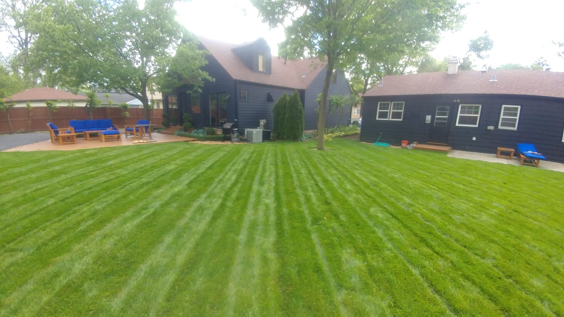 A backyard with neatly mowed grass in stripes, two dark houses with red roofs, patio furniture, trees, and a wooden fence bordering the area.