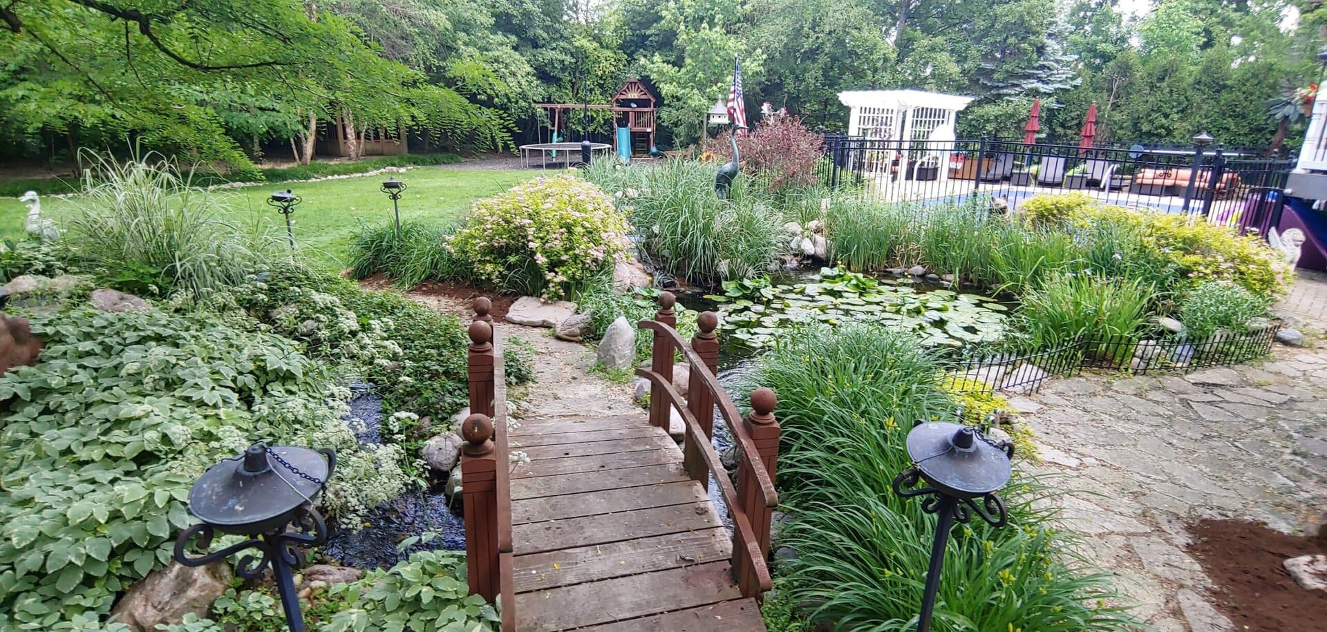 A small wooden bridge crosses a lush garden pond with lily pads, surrounded by greenery, flowering bushes, and decorative lanterns, leading to a yard with a gazebo and fenced pool area.