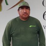 A man in a green Sweeneys shirt and cap stands in front of a wall with partial text and graphics. He has a mustache and is looking at the camera with a neutral expression.