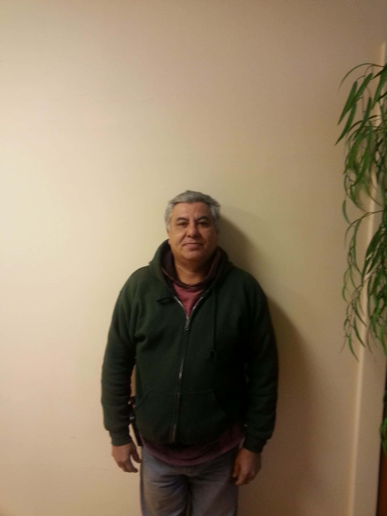 A man with short gray hair, wearing a green zip-up hoodie and maroon shirt, stands against a beige wall. A leafy green plant is visible on the right side of the image.