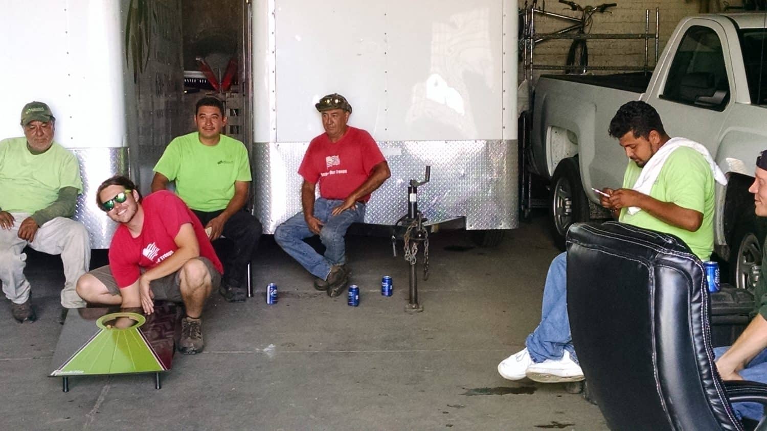 Six men sit or relax near a white trailer and pickup truck in a garage. Some wear green or red shirts. One plays cornhole, another checks his phone, and soda cans are on the ground.