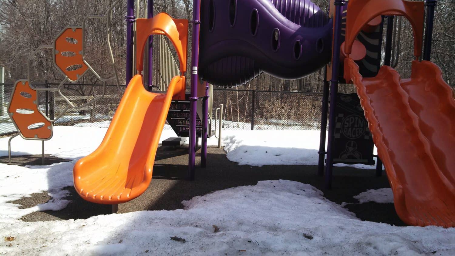 Two orange slides and climbing walls on a purple playground structure, surrounded by snow patches and bare trees in the background.