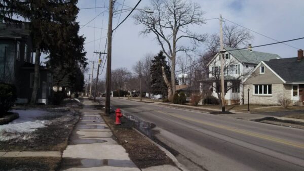 A quiet suburban street with bare trees, houses, a red fire hydrant, and patches of melting snow on the sidewalk and grass.