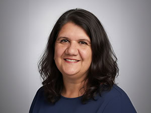 Woman with shoulder-length dark hair wearing a navy blue top, smiling at the camera in front of a plain light gray background.