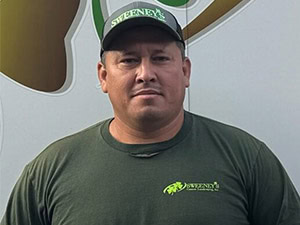 Man wearing a dark green Sweeneys t-shirt and cap stands in front of a wall with a green and brown logo. He looks directly at the camera with a neutral expression.