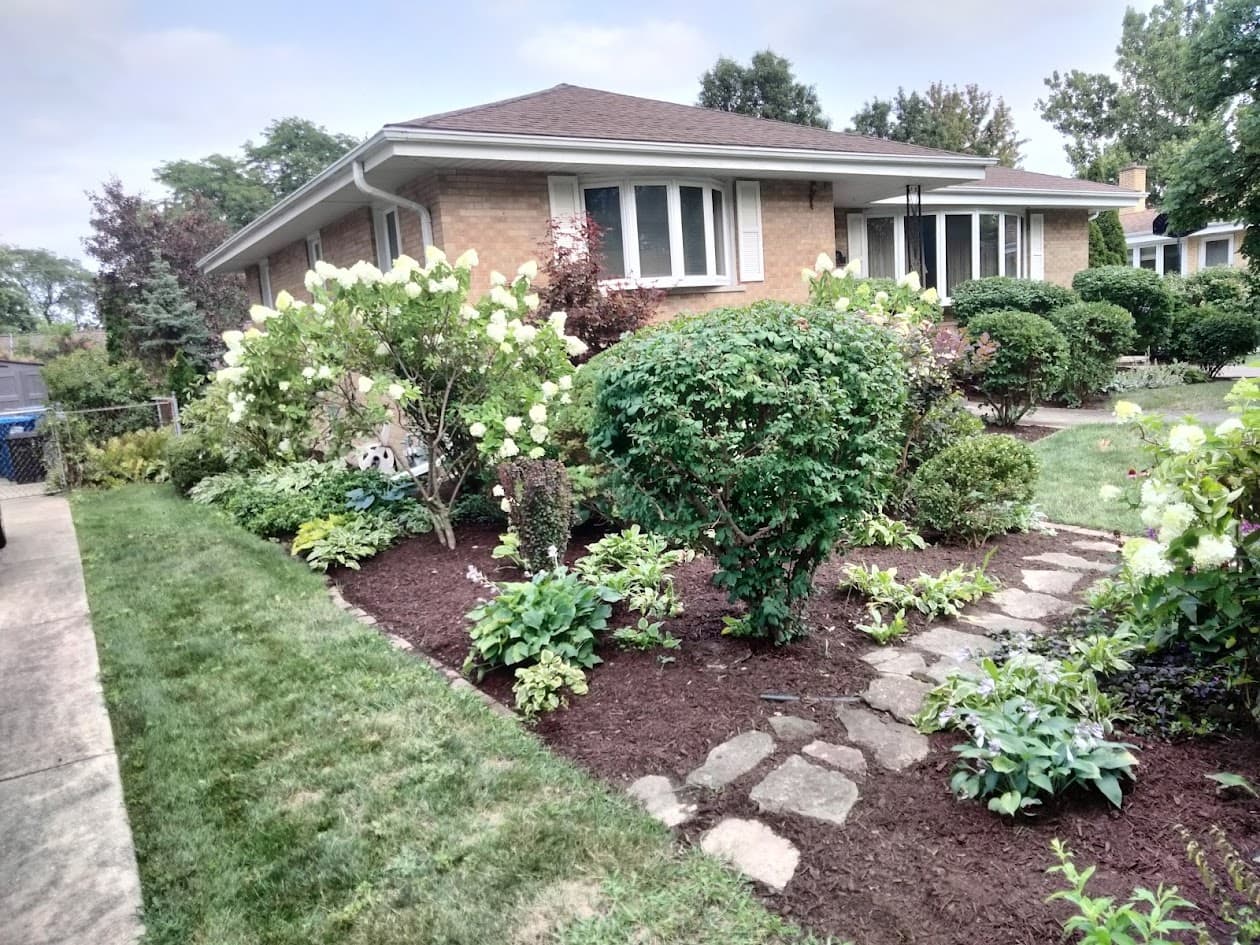 A neatly landscaped front yard with bushes, flowering plants, and stone path leading to a brick house with white-trimmed windows. The lawn is well-maintained and there are trees in the background.
