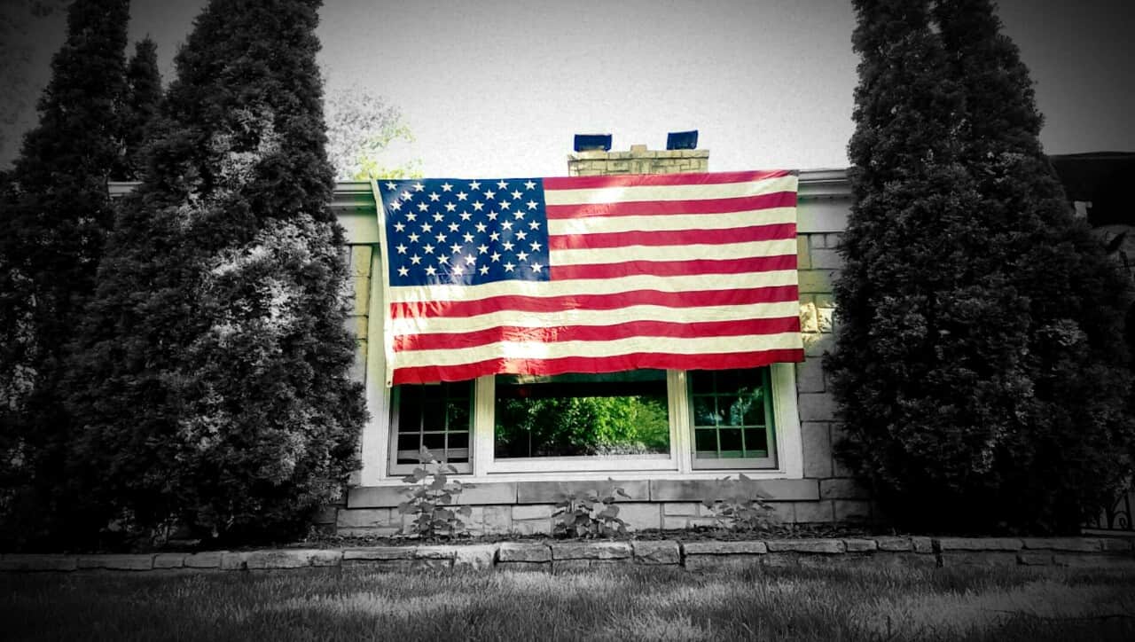 A large American flag hangs over a window on a houses front facade, flanked by two tall evergreen trees, with grass and shrubs in the foreground.