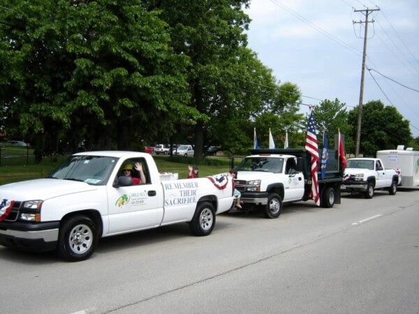 Three white trucks decorated with flags and bunting drive down a street in a parade. One truck has a sign reading Remember Their Sacrifice. People are riding in or driving the vehicles. Trees and power lines are visible.