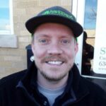 A smiling man with a light beard and mustache wears a black jacket and a black cap, standing in front of a building with a sign partially visible in the background.