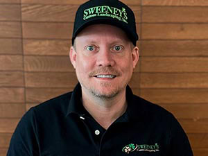 A man wearing a black Sweeneys Landscaping, Inc. hat and black polo shirt smiles at the camera. He is sitting in front of a wooden wall.