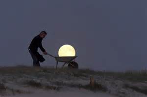 A person pushes a wheelbarrow on a grassy hill at dusk, positioned so the full moon appears to be sitting inside the wheelbarrow.
