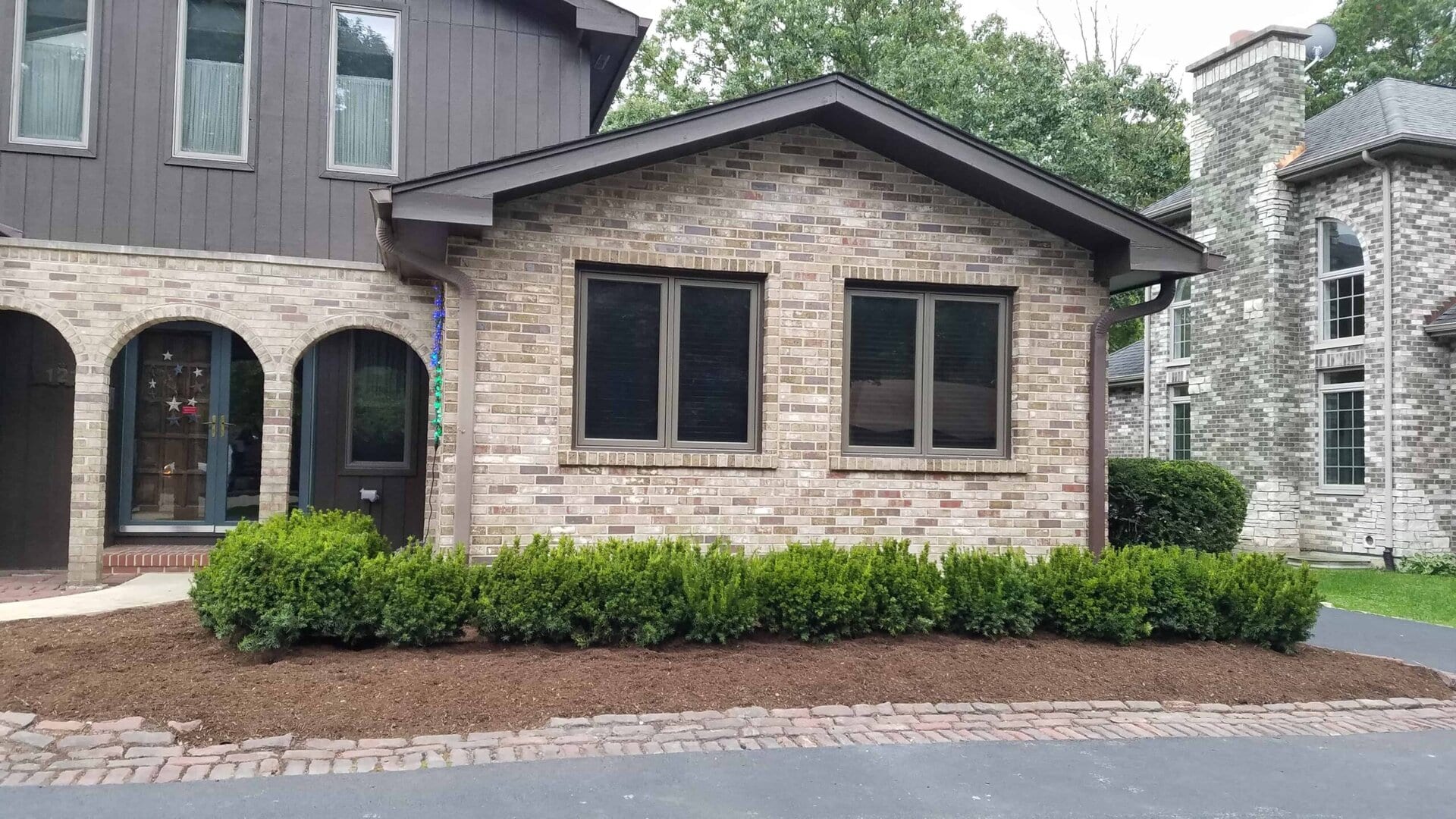 Single-story extension with three large windows and light brick exterior, bordered by green shrubs and mulch, connected to a larger dark brown house, with neighboring homes and trees in the background.