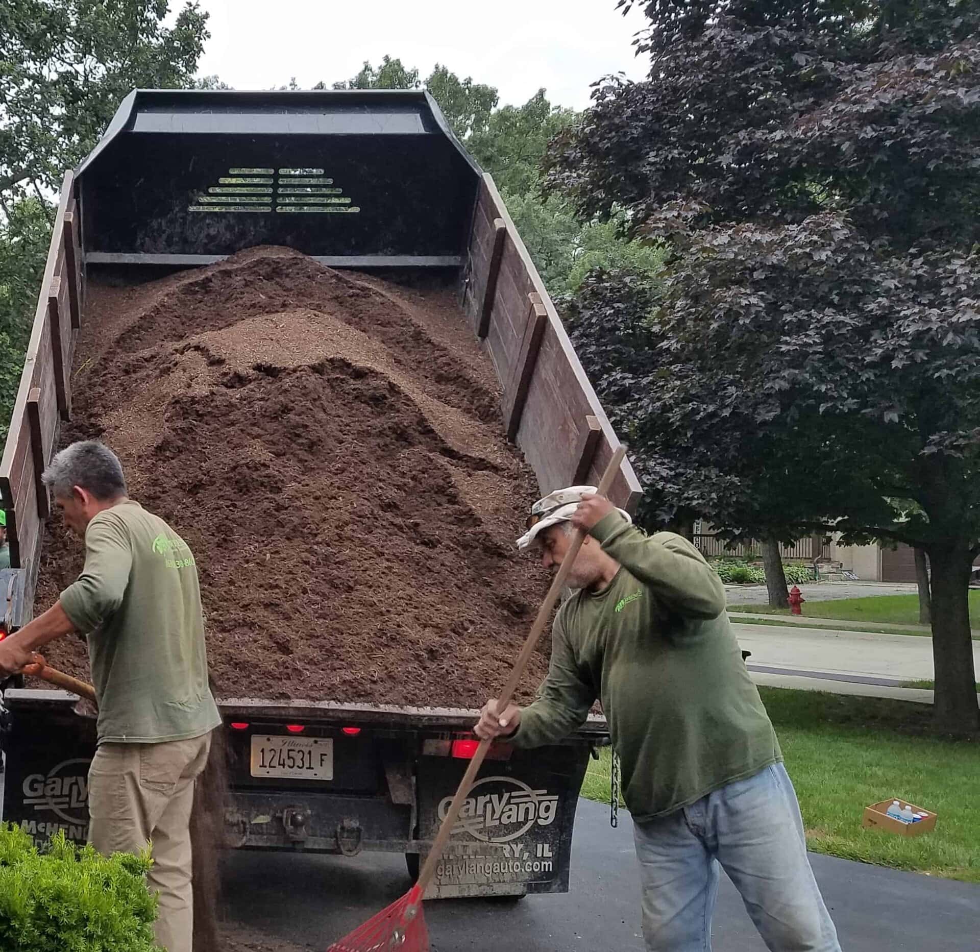 Two workers shovel dirt from the back of a dump truck onto a driveway in a suburban neighborhood. One is holding a shovel while the other is inside the truck bed. Trees and houses are visible in the background.