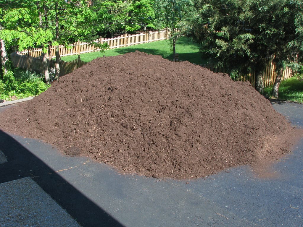 A large pile of brown mulch sits on a driveway, surrounded by green trees, grass, and a wooden fence in the background.