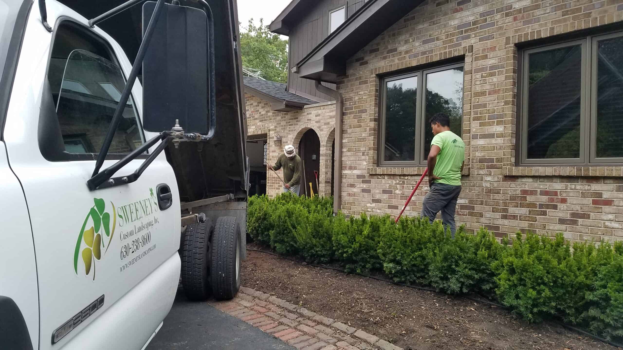 Two people work on landscaping near a brick house, using rakes to tend soil by a row of shrubs. A white Sweeper truck is parked nearby with its door open, partially visible in the foreground.