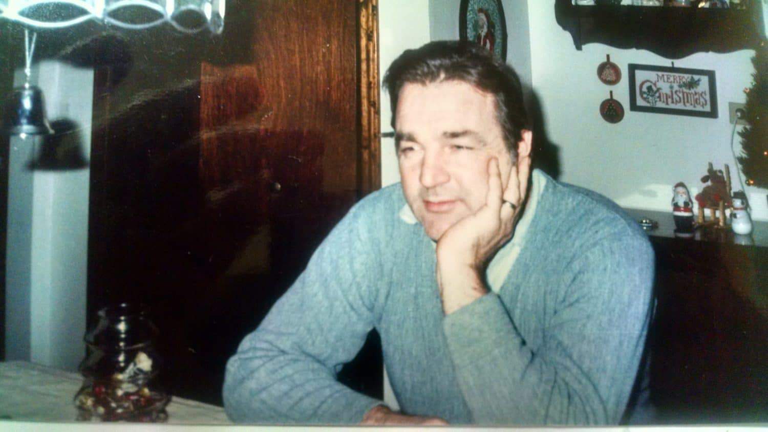 A man in a light blue sweater sits at a table with his head resting on his hand. There are Christmas decorations in the background, including a Merry Christmas sign on the wall.