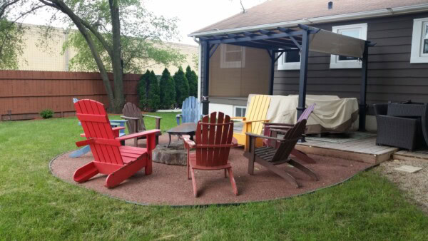 Six colorful Adirondack chairs arranged around a fire pit in a backyard with grass, a pergola-covered patio, and a brown fence in the background.