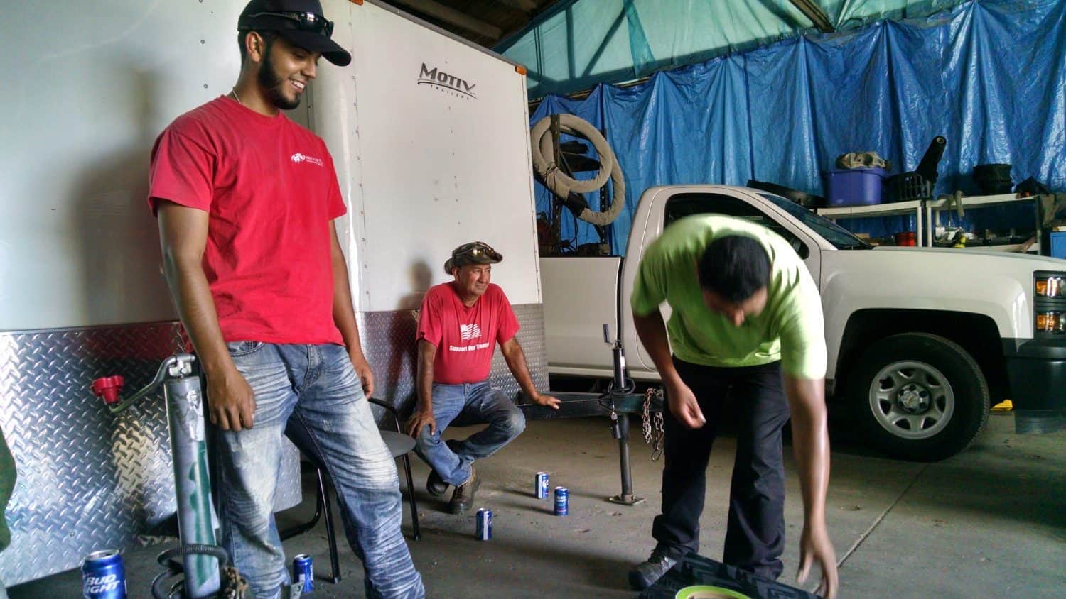 Three men are in a garage near a white truck and a trailer. One man in red stands smiling, another sits, and a third in green bends over. Several cans are on the floor, and tools and equipment are visible around them.