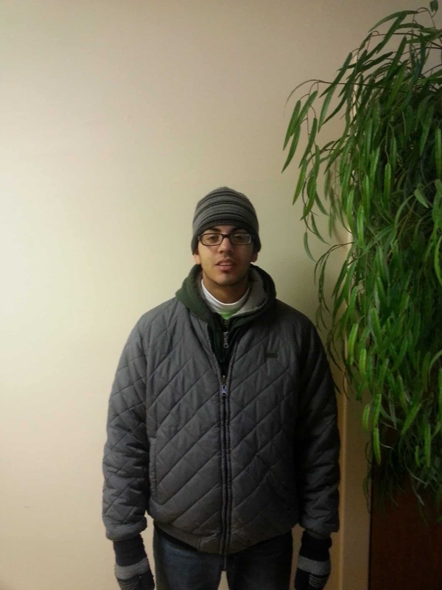 A person wearing glasses, a striped beanie, a gray quilted jacket, gloves, and jeans stands indoors next to a tall green plant against a plain light-colored wall.