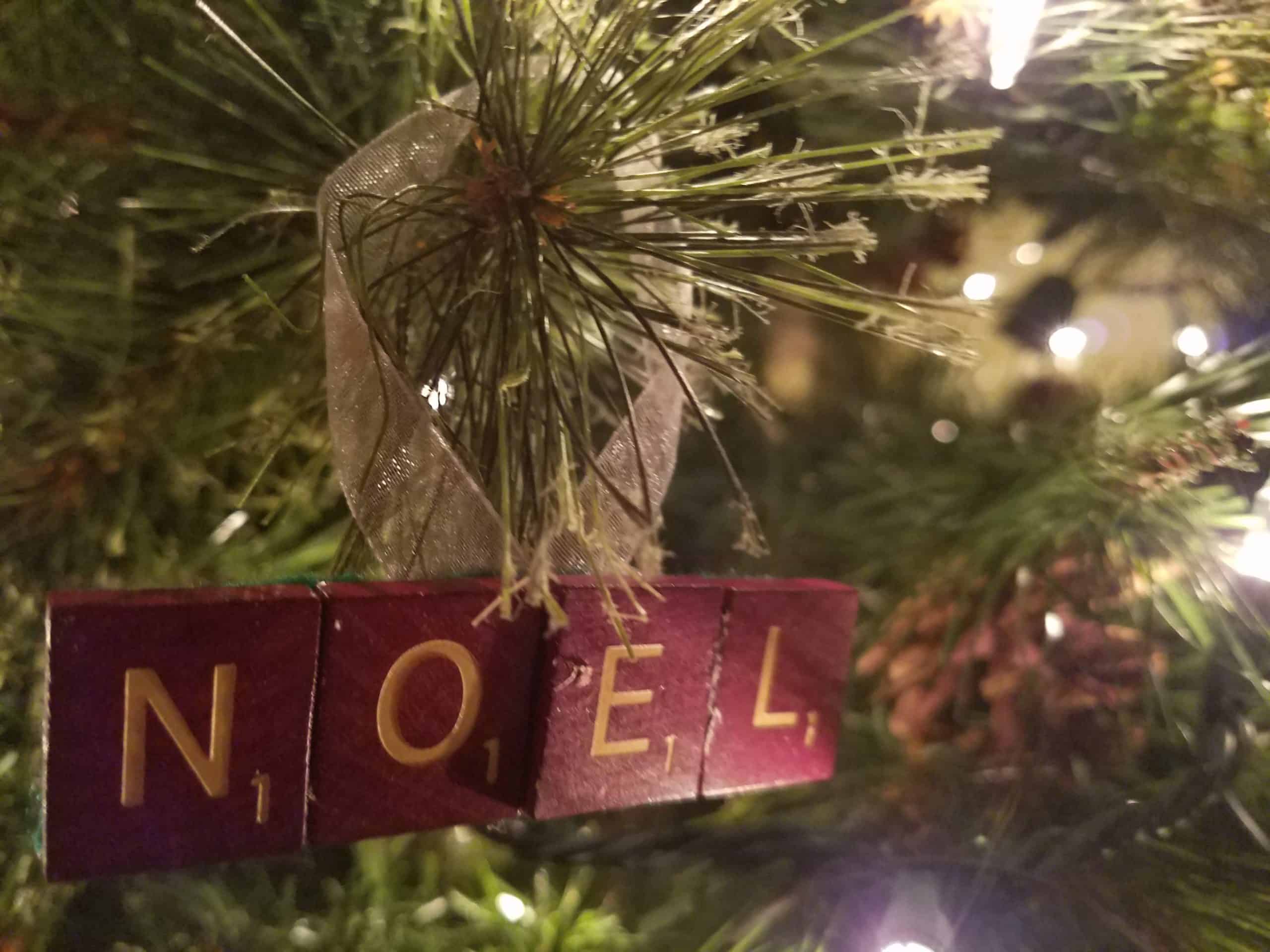 Close-up of a Christmas tree branch with a ribbon and four Scrabble tiles spelling NOEL as an ornament, surrounded by festive lights and pine needles.