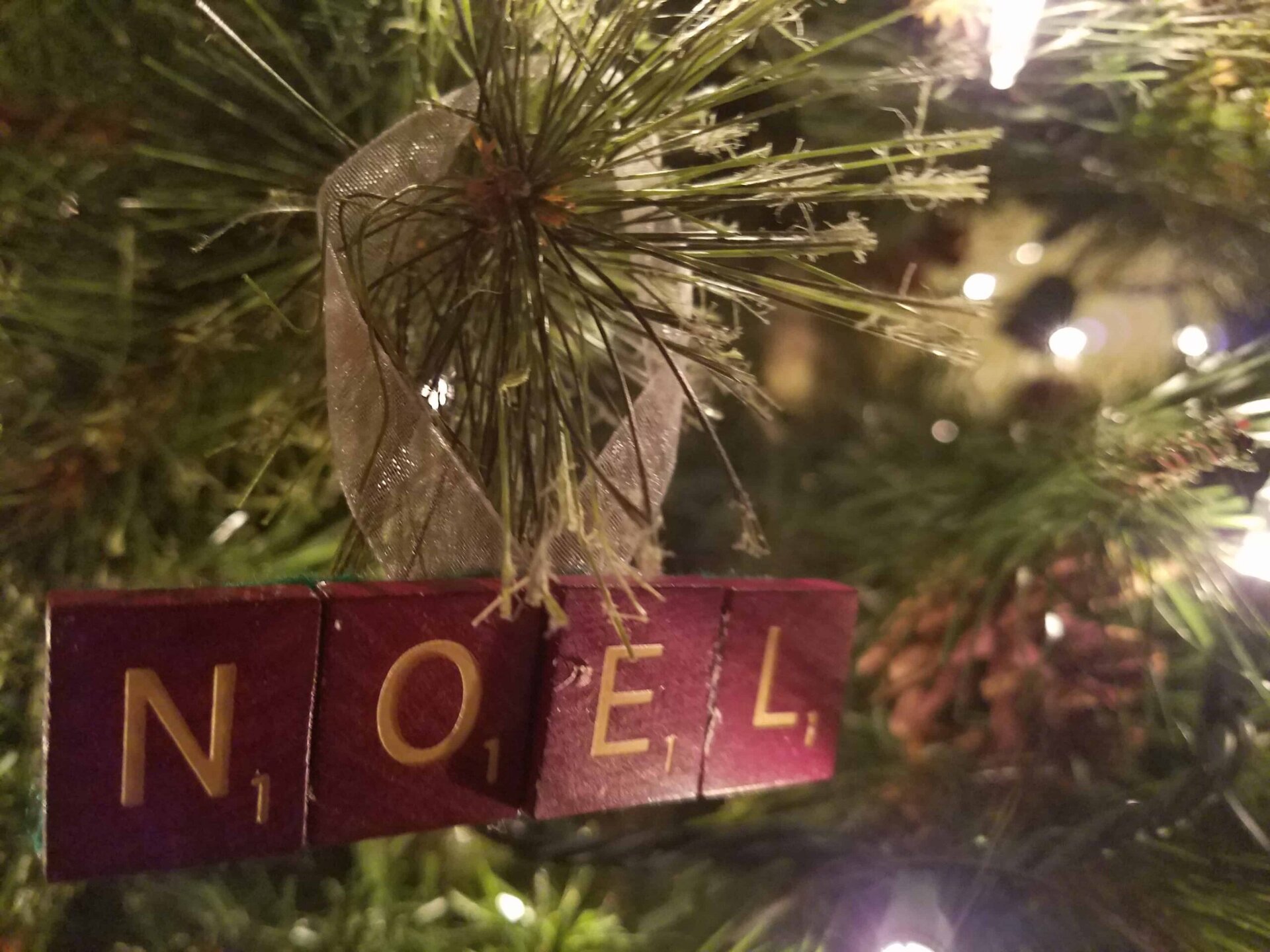 Close-up of a Christmas tree branch with a ribbon and Scrabble tiles spelling NOEL hanging as an ornament, surrounded by festive lights.