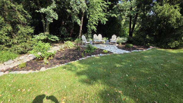 Four white Adirondack chairs are arranged on a stone patio surrounded by plants and trees, with a grassy lawn in the foreground and partial shadows visible.
