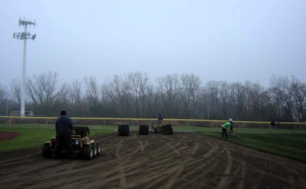 Three people work on a foggy baseball field: one operates a machine on dirt, while two others lay or adjust sod near the outfield fence. Bare trees line the background.