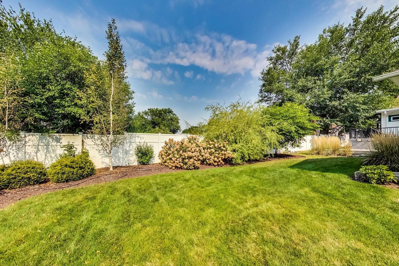 A backyard with green grass, shrubs, and trees, bordered by a white fence under a blue sky with scattered clouds.