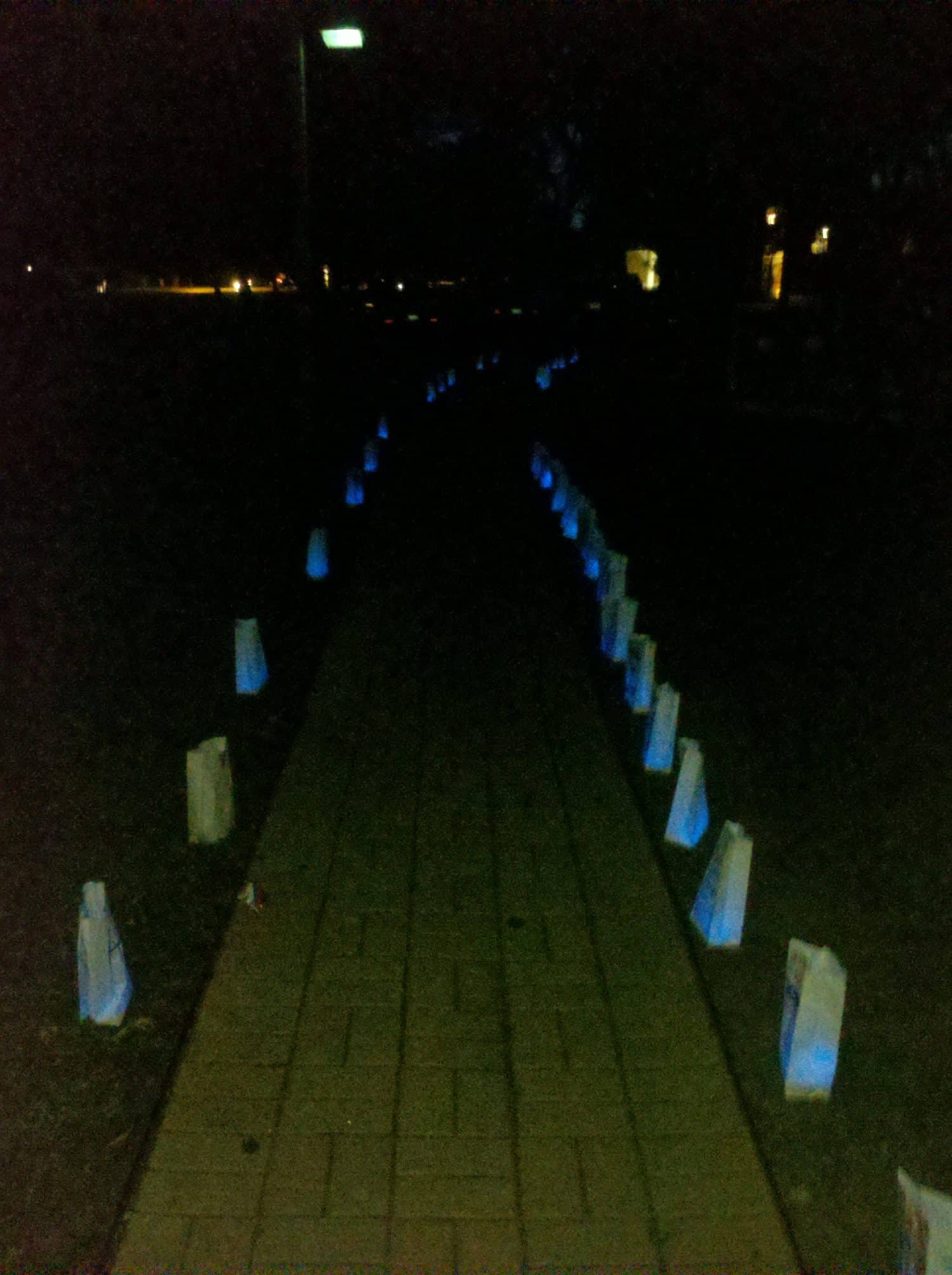 A paved path at night is lined with glowing blue luminaries in white paper bags on both sides, creating a lit walkway through the darkness.