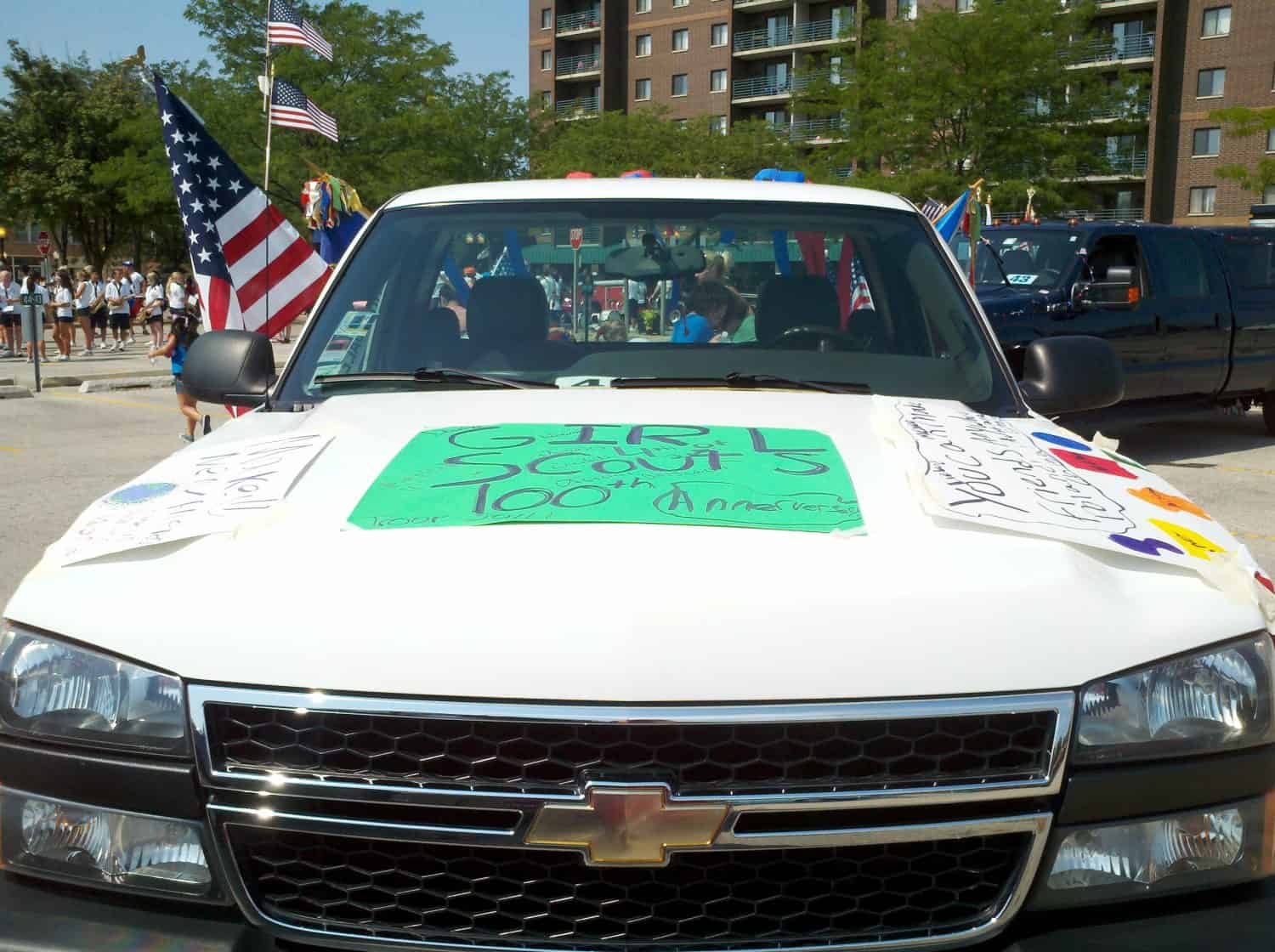 A white Chevrolet truck decorated with signs reading Girl Scouts 100+ Anniversary is parked outdoors during a parade. American flags and people are visible in the background.