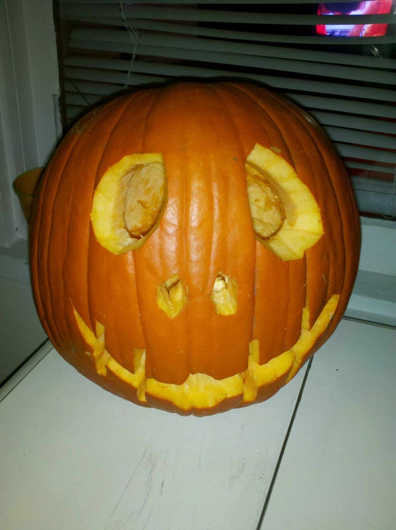 A carved pumpkin with an uneven, wide smile and oval eyes sits on a white table by a window with blinds.
