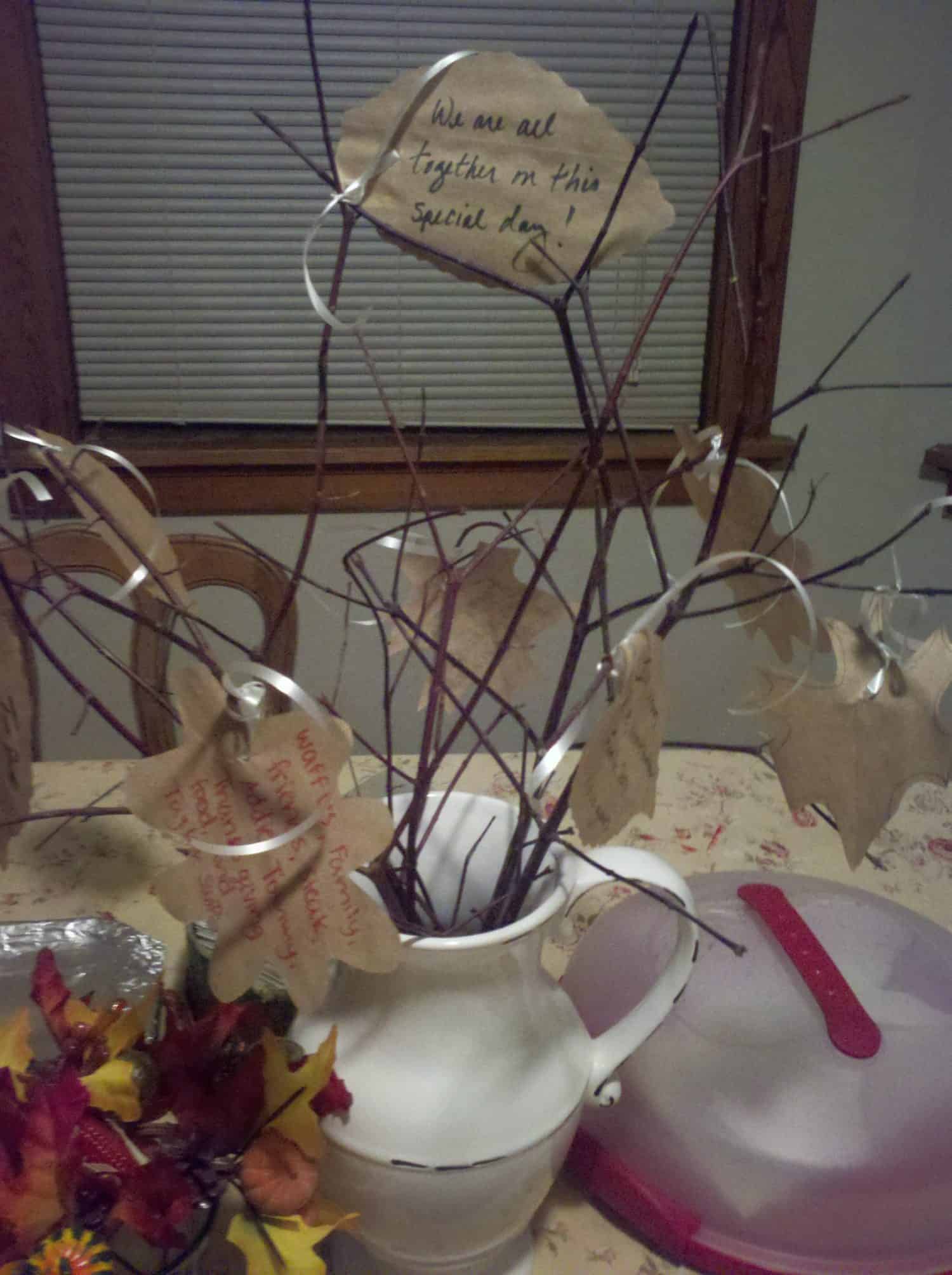 A white pitcher holds bare branches with handwritten paper leaves attached. The centerpiece sits on a table with fall decorations and a covered dish nearby. One leaf reads, We are all together on this special day!.