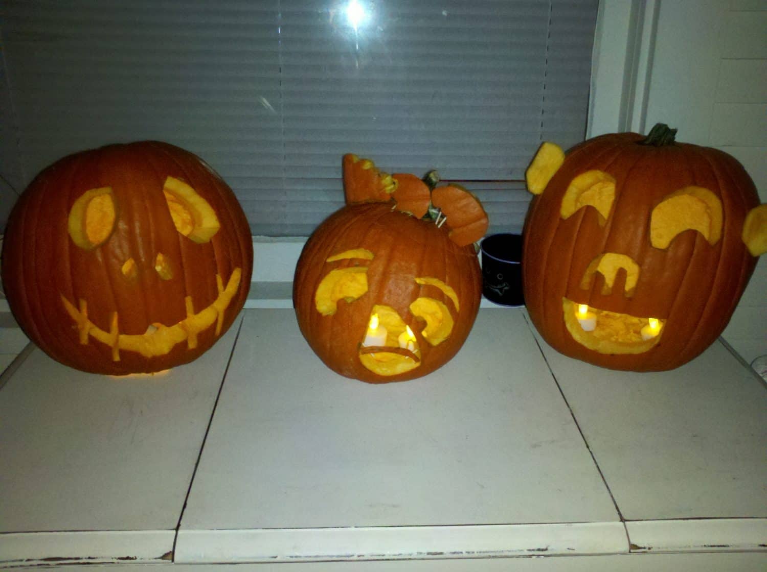 Three carved pumpkins with lit candles inside sit on a white table. Each pumpkin has a different silly face: one with an awkward smile, one with a distressed look, and one with bear-like ears and a happy face.