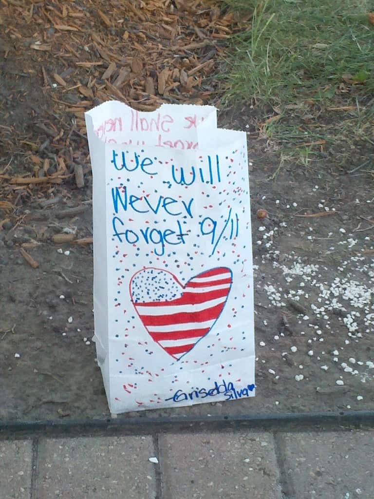 A decorated white paper bag with an American flag heart and the handwritten words We will never forget 9/11, placed on the ground near grass.