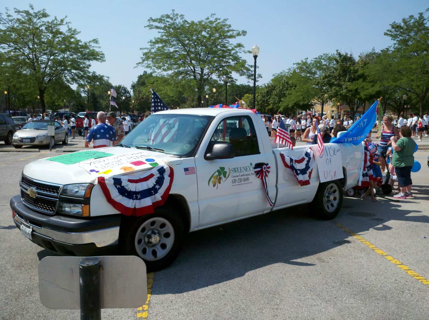 Pic #2 A white pickup truck decorated with international flags and patriotic banners is parked at an outdoor event. People nearby are holding American flags, and trees line the background.