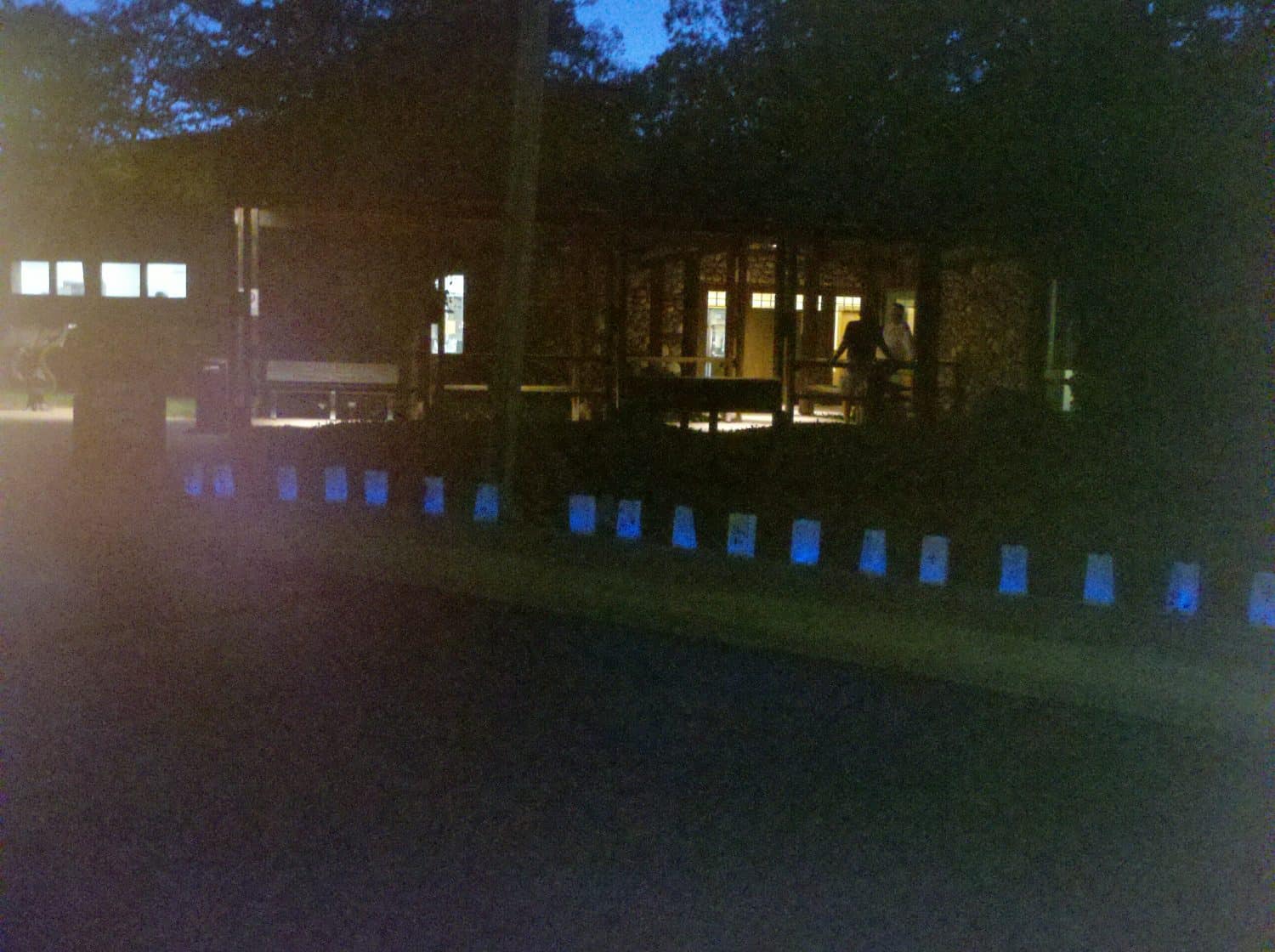 A row of glowing lantern bags lines a sidewalk at night, in front of a pavilion with lights on inside and surrounded by trees.