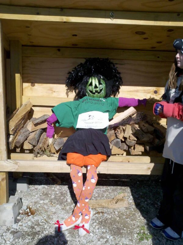 Scarecrow with a painted green pumpkin head, black wig, green shirt, orange skirt, and patterned tights sits on a bench of stacked firewood. A person stands nearby, partially out of frame.