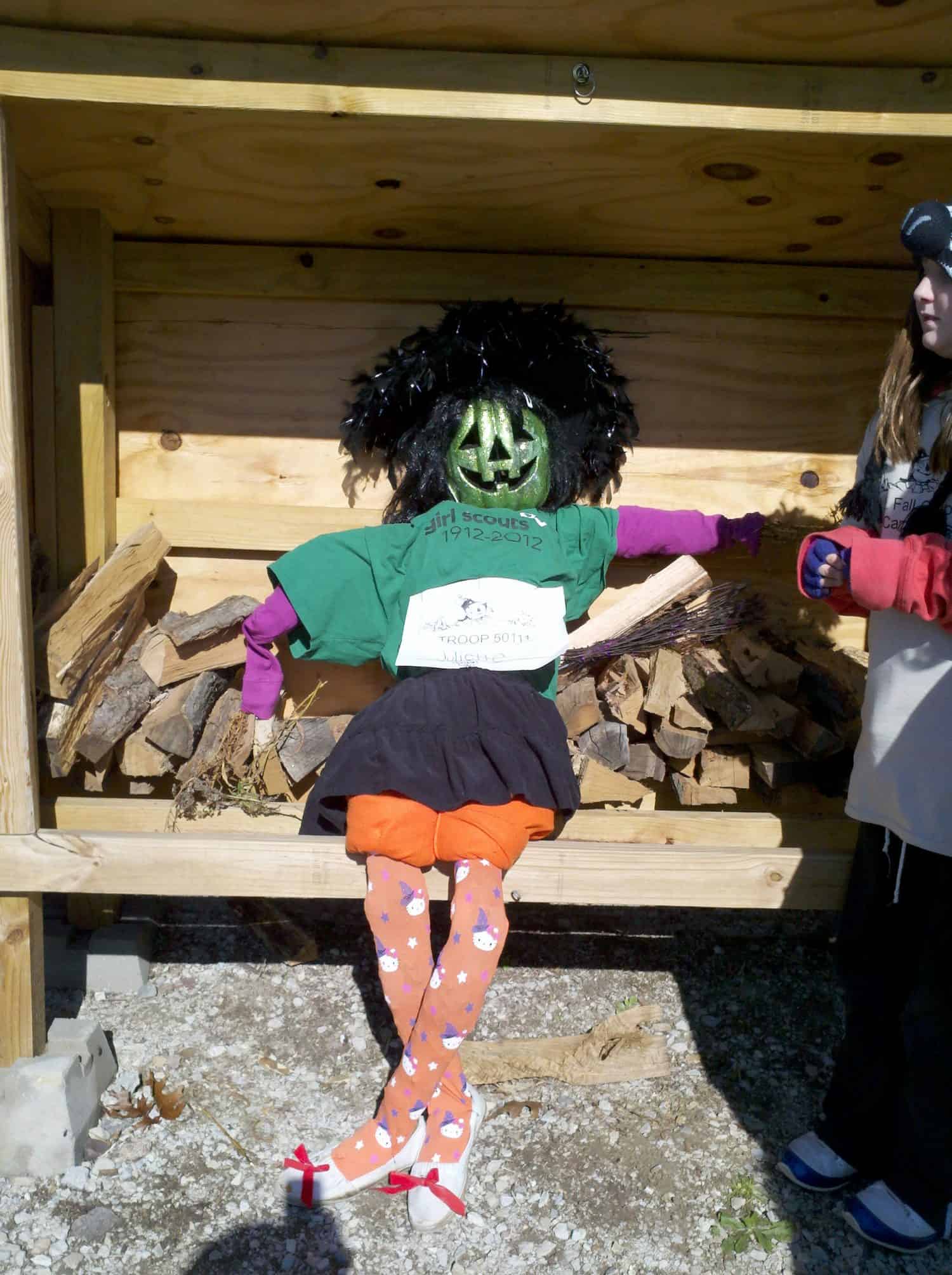 Pic #32 A scarecrow with a green pumpkin head, green shirt, black skirt, orange tights, and a black wig sits on a woodpile. A person stands nearby, partially visible, wearing a costume with gloves.