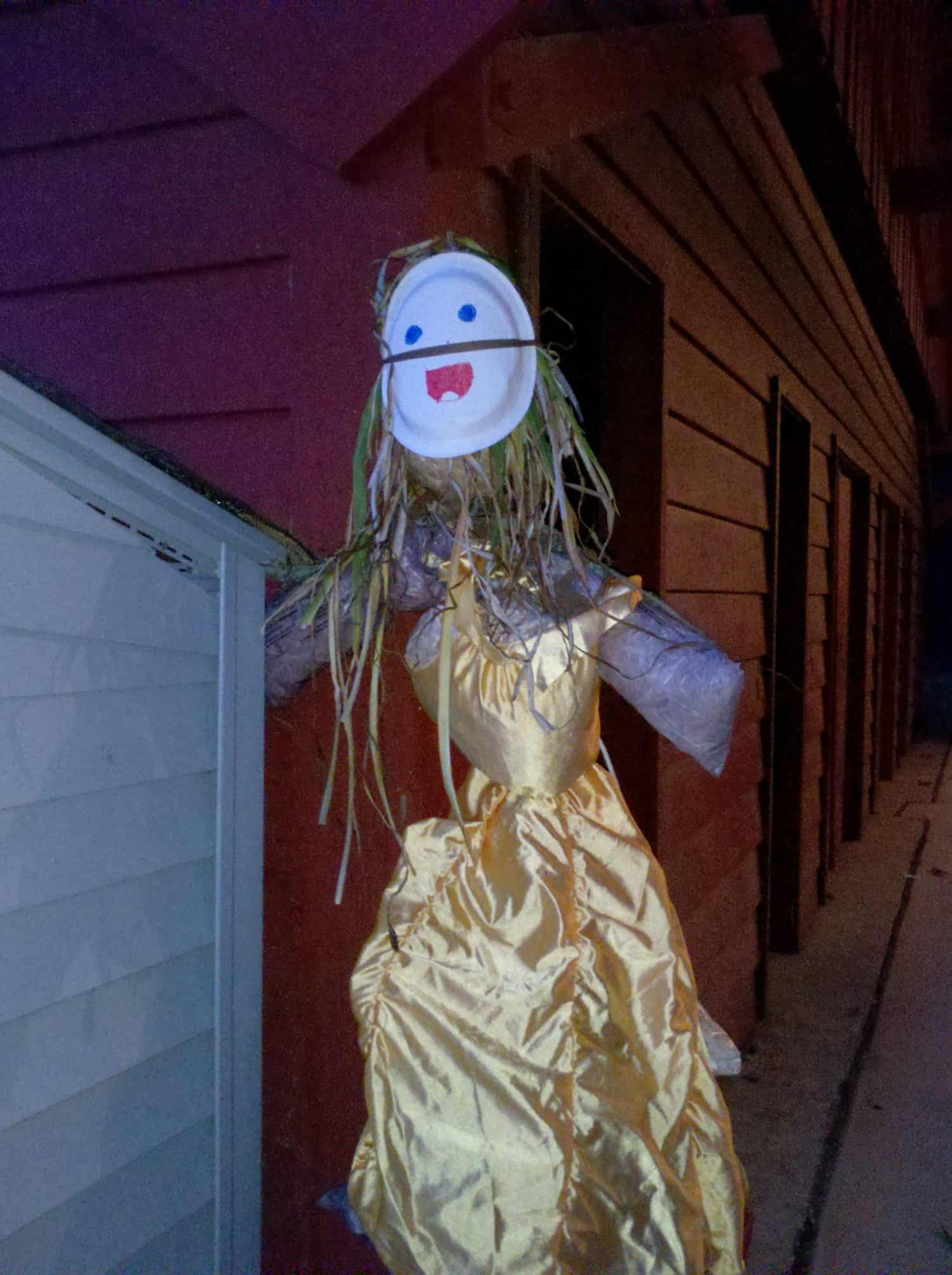 Pic #47 A scarecrow with a paper plate face, yarn hair, and a yellow dress stands near a building at night.