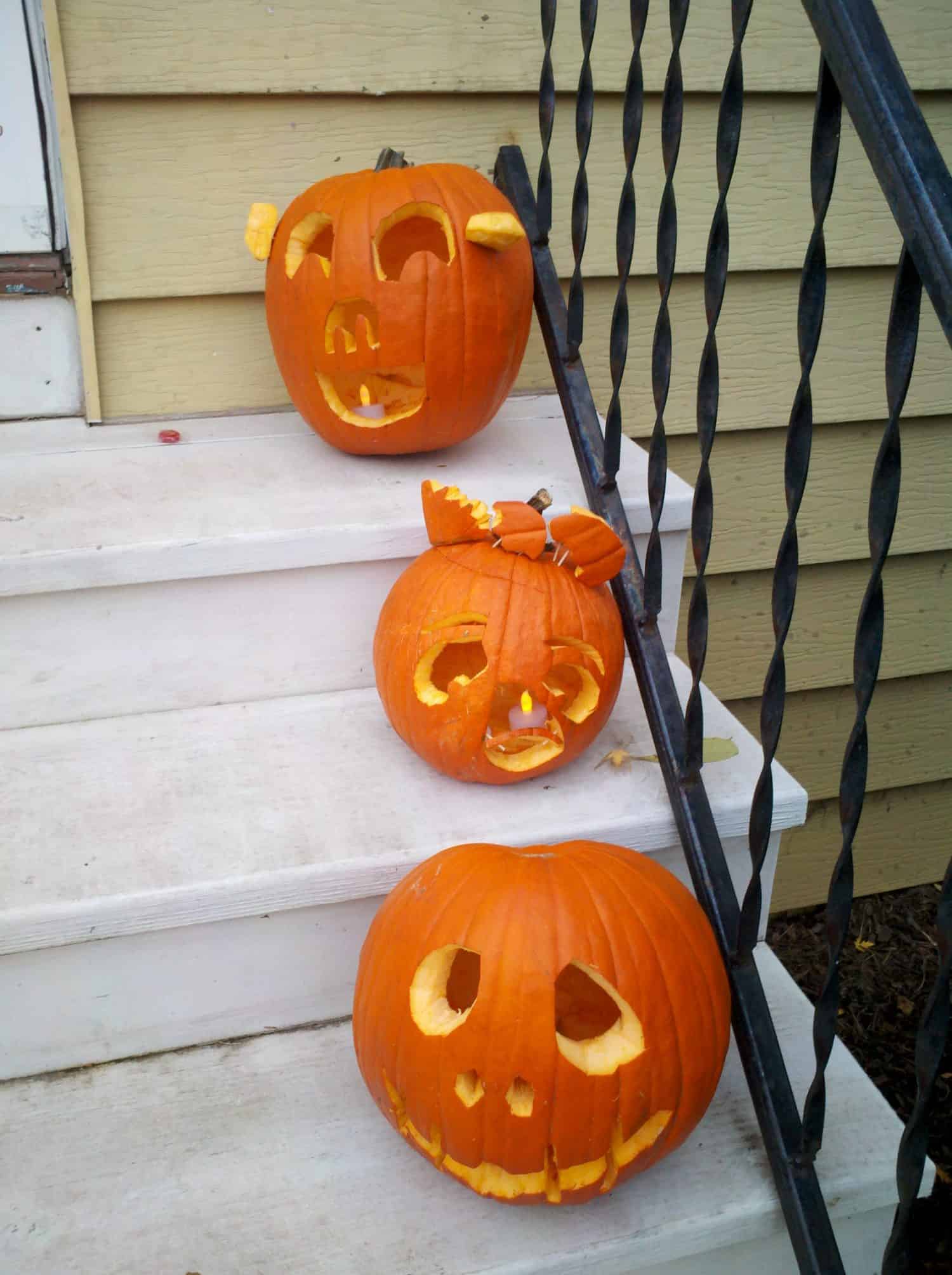 Three carved pumpkins with various facial expressions sit on white steps next to a black iron railing. The pumpkins have different designs and some broken or missing pieces.