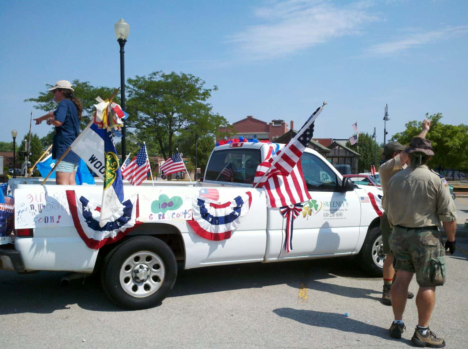 Pic #5 A white pickup truck decorated with American flags, bunting, and signs is parked. Several people stand nearby, some in hats and shorts, as part of a parade or celebration on a sunny day.