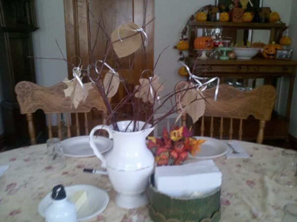 A white pitcher with bare branches and paper leaves sits on a dining table with a floral cloth. The table is set for a meal, and pumpkin decorations are visible in the background on a wooden sideboard.