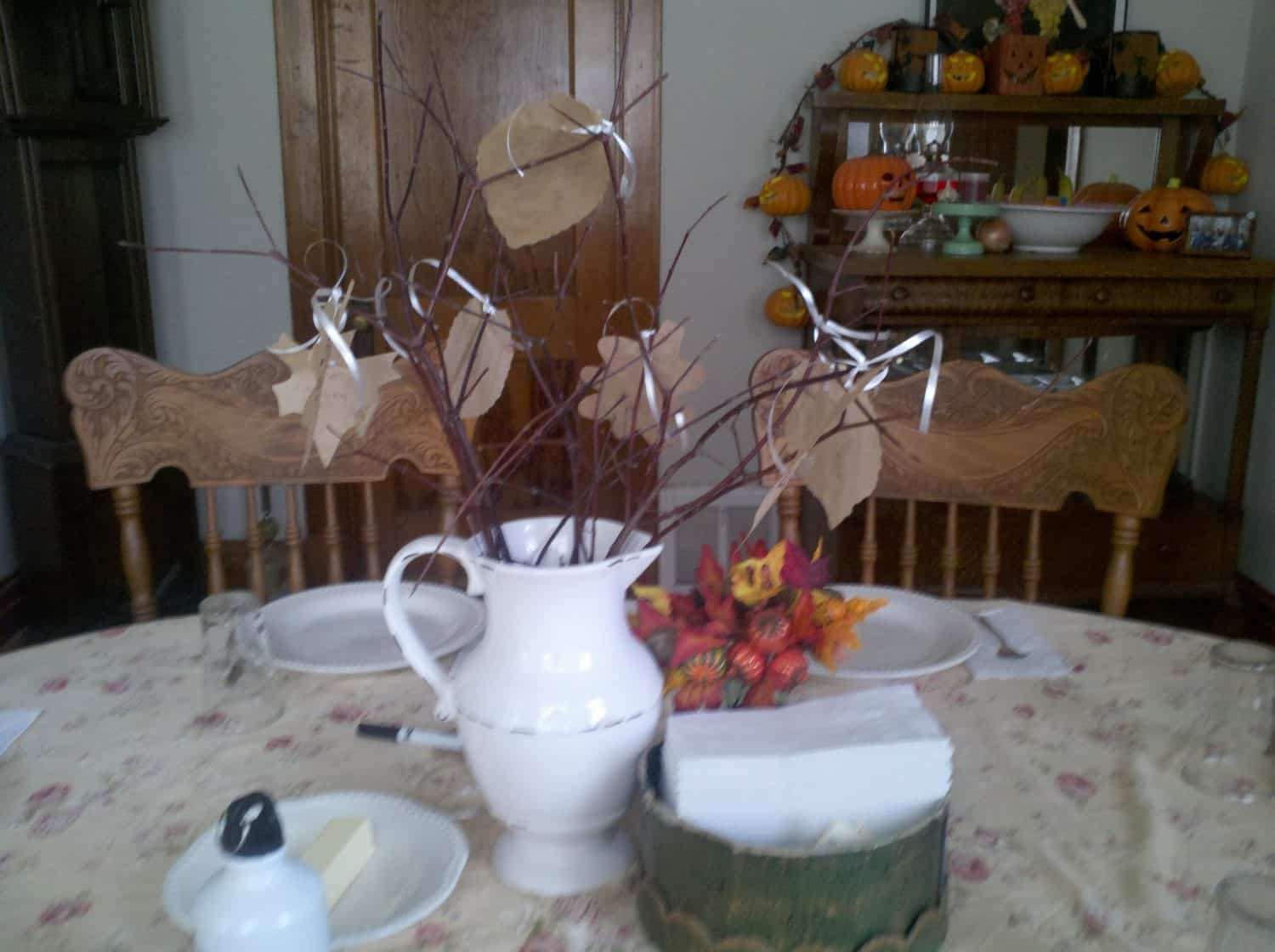 A white pitcher with bare branches and paper leaves sits on a dining table with a floral cloth. The table is set for a meal, and pumpkin decorations are visible in the background on a wooden sideboard.