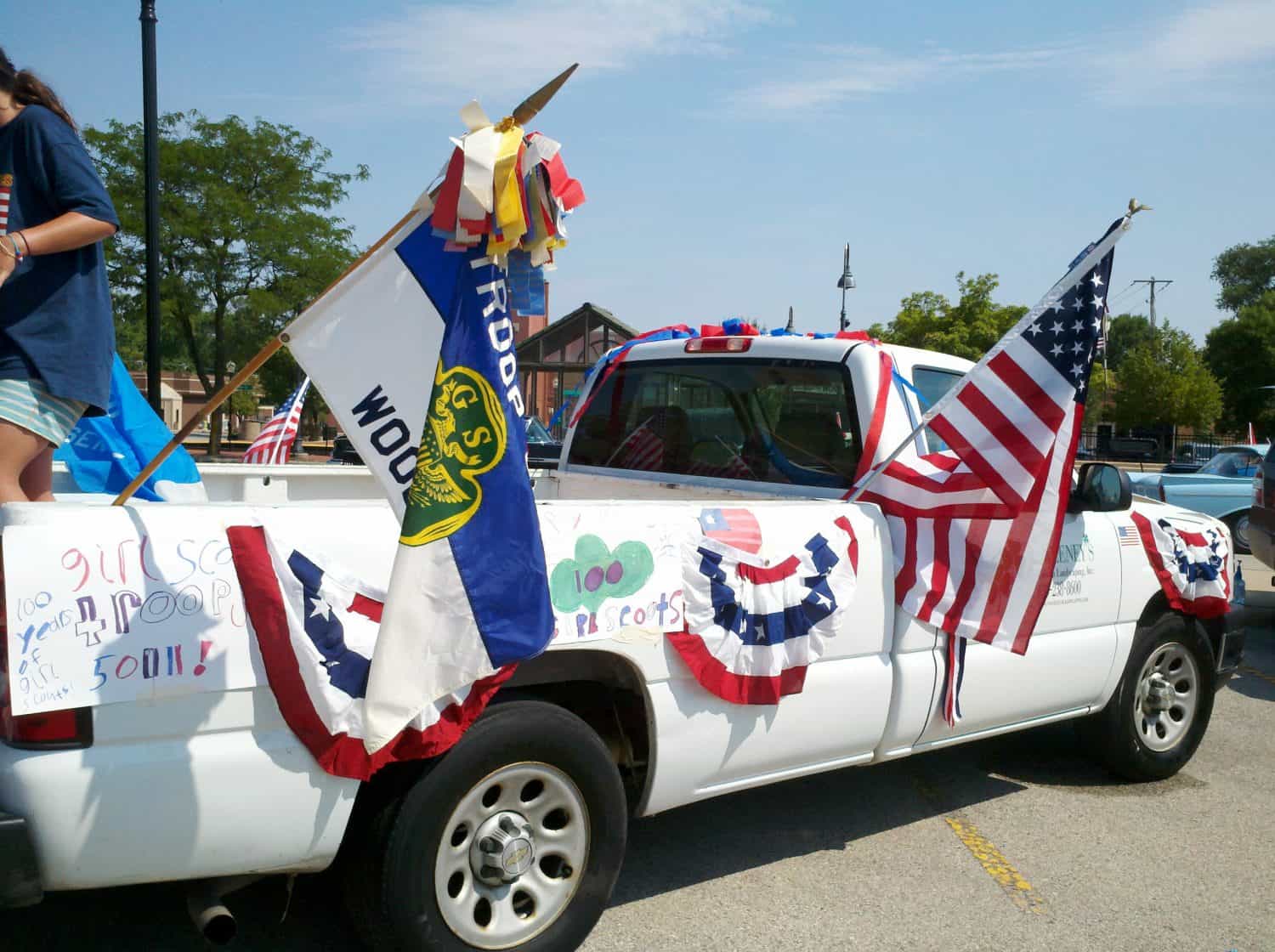 A white pickup truck decorated with American flags, banners, and handmade signs, including Girl Scout references, parked outdoors on a sunny day for a parade or celebration.
