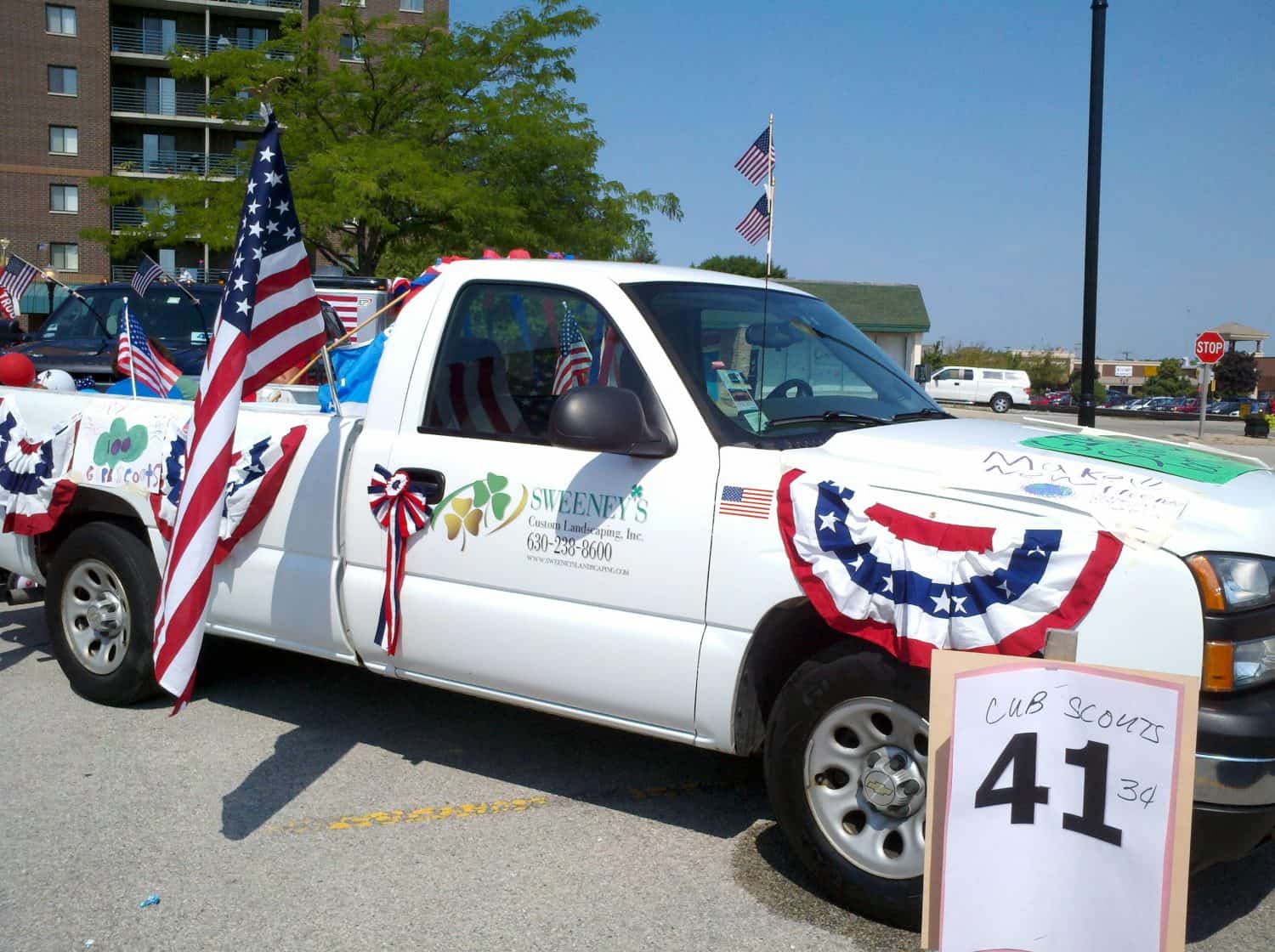 A white pickup truck decorated with American flags and red, white, and blue bunting is parked in a lot. A sign in front reads Cub Scouts 41 3rd. Other flags and parade decorations are visible.